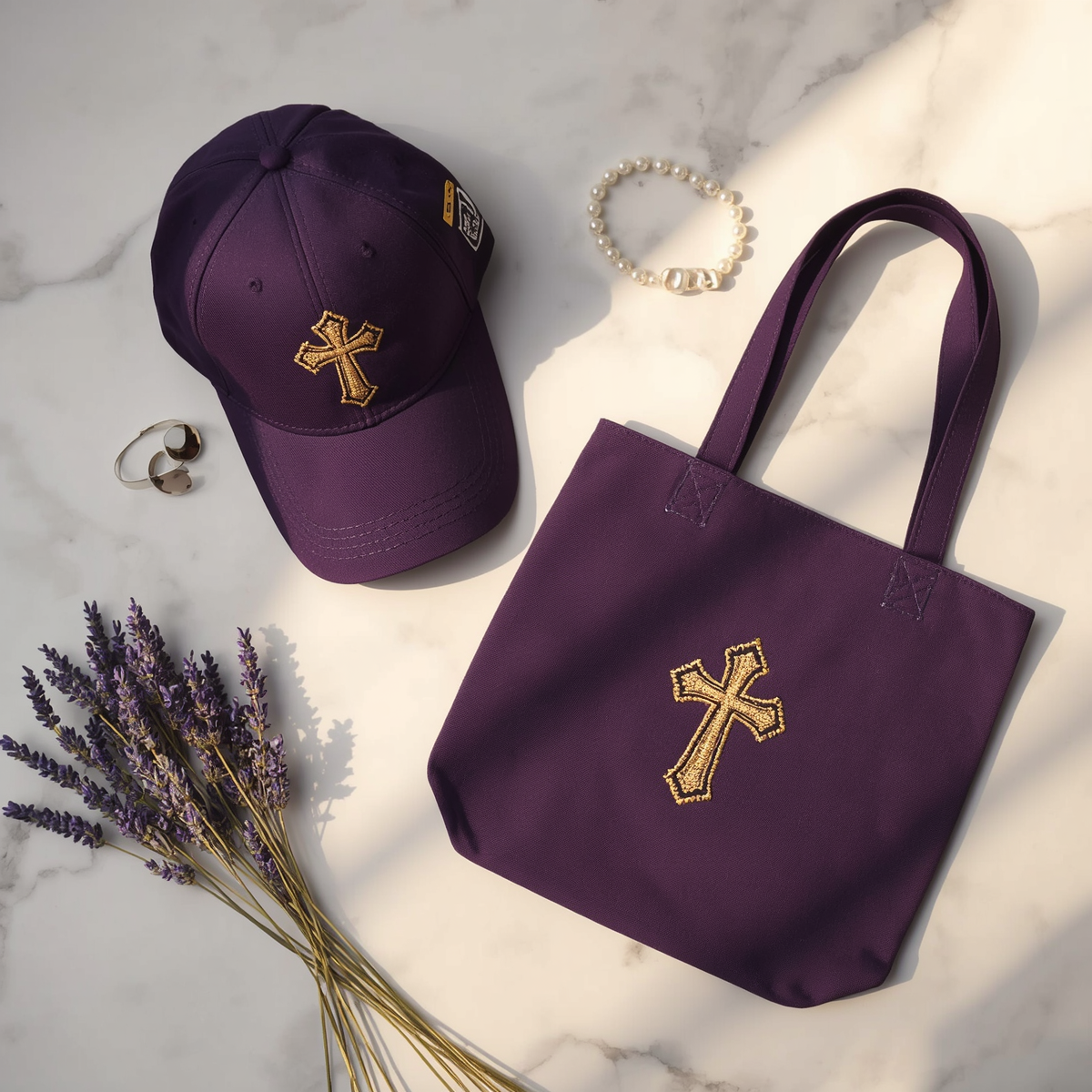 A deep purple embroidered baseball cap with a gold cross design, a matching tote bag, and pearl accessories arranged on a white marble surface with dried lavender flowers.