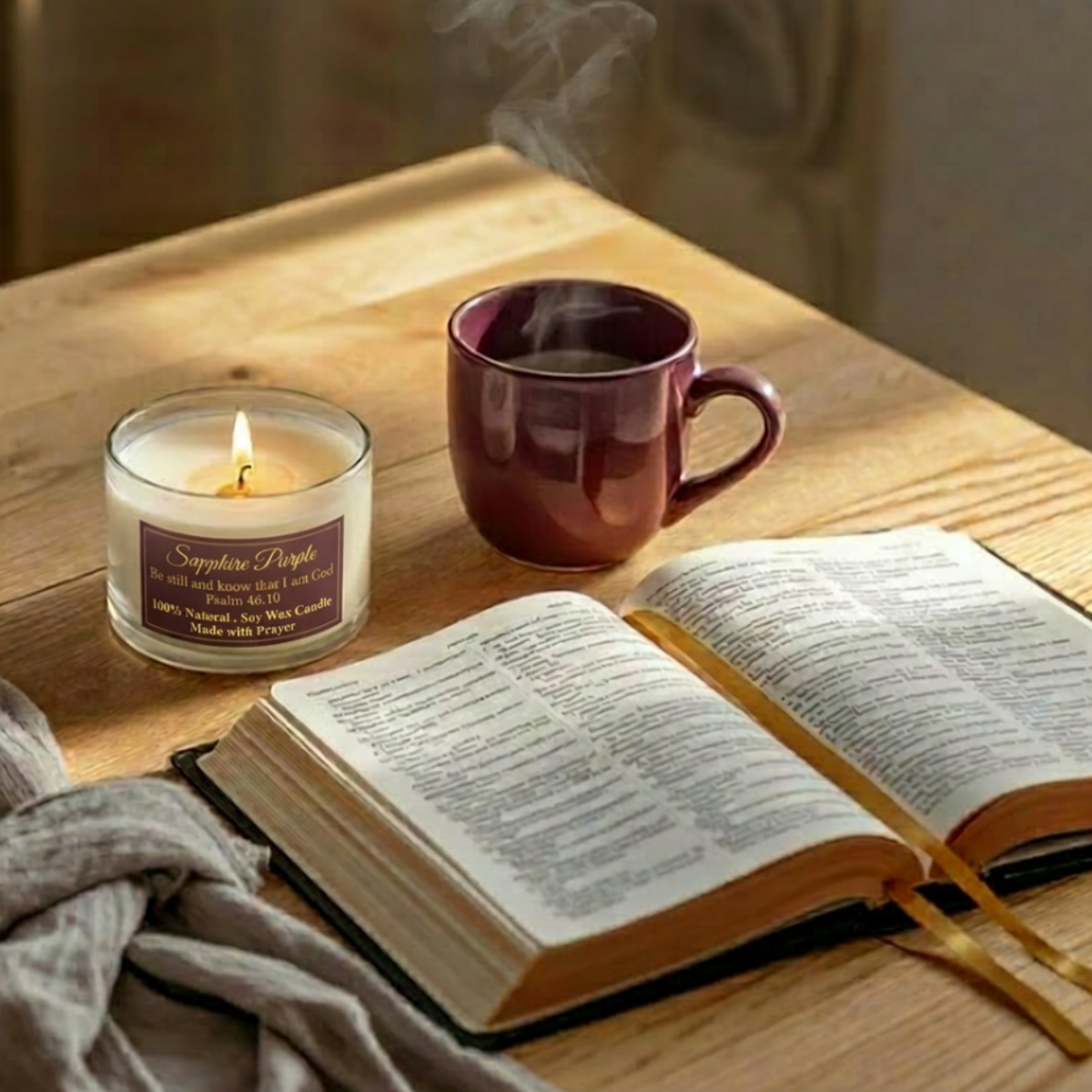 A warm cozy styled home featuring a lit white soy candle with a deep purple label, a deep purple ceramic mug with steam rising, an open Bible with a gold ribbon bookmark.